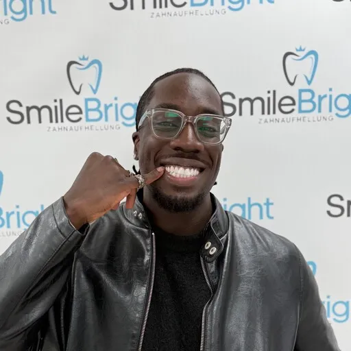 Ein lächelnder Mann in einer schwarzen Lederjacke und Brille steht vor einer Wand mit dem Logo "Smile Bright Zahnaufhellung". Er zeigt mit dem Finger auf seine hellen Zähne.