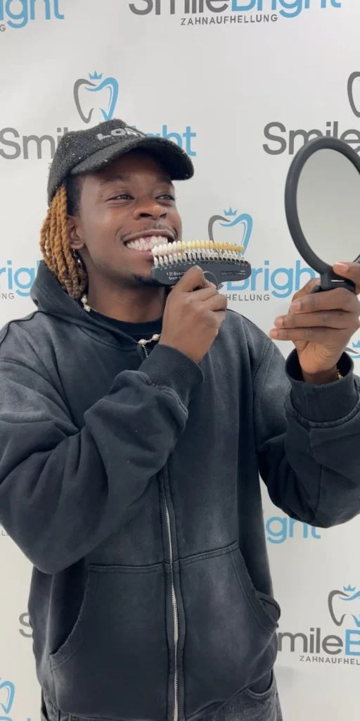 Ein glücklicher Kunde betrachtet sein strahlend weißes Lächeln in einem Handspiegel und vergleicht das Ergebnis mit einer Zahnfarbenskala, während er vor der Smile Bright Logo-Wand in Düsseldorf steht.