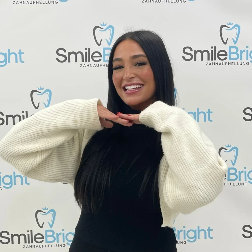 Glückliche Kundin mit langen dunklen Haaren rahmt ihr strahlend weißes Lächeln stolz mit den Händen ein. Sie posiert nach ihrer Behandlung vor der Smile Bright Zahnaufhellung Logo-Wand in Düsseldorf.