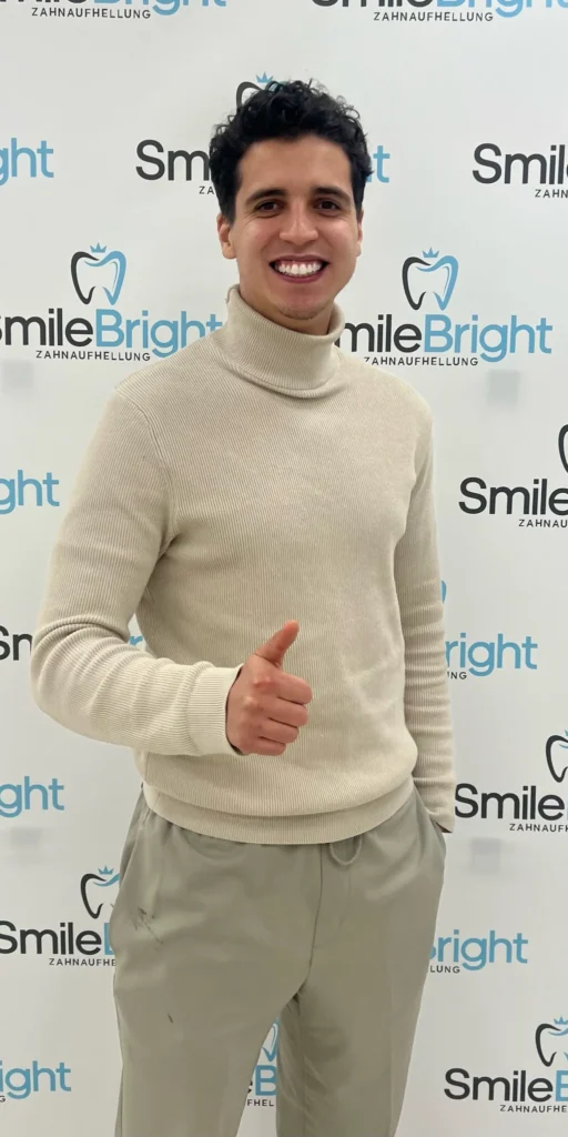 Zufriedener junger Mann im beigen Rollkragenpullover mit einem strahlend weißen Lächeln, der nach seiner Behandlung vor der Smile Bright Zahnaufhellung Logo-Wand in Düsseldorf einen Daumen nach oben zeigt.