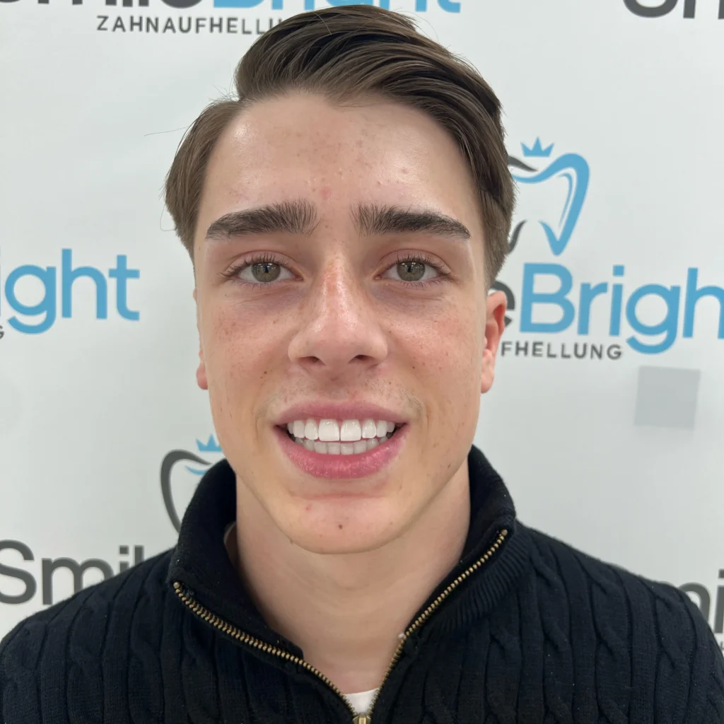 Zufriedener Kunde mit einem confidenten, strahlend weißen Lächeln nach einem kosmetischen Bleaching vor der Smile Bright Zahnaufhellung Düsseldorf Logo-Wand.