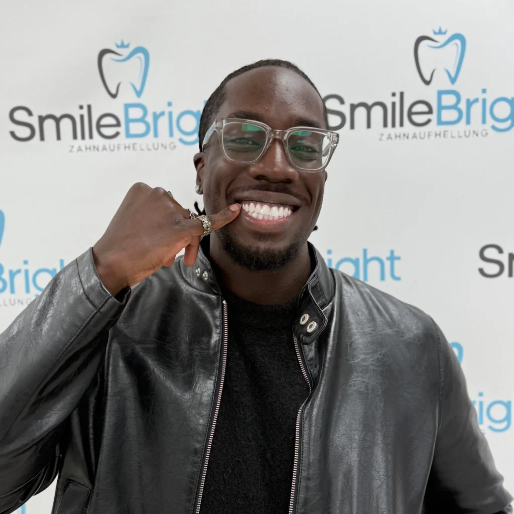 Glücklicher Schwarzer Mann mit klarem Brillengestell und schwarzer Lederjacke, der eine "Ruf mich an"-Geste macht und breit lächelt, vor dem Smile Bright Zahnaufhellung Logo-Hintergrund in Düsseldorf.