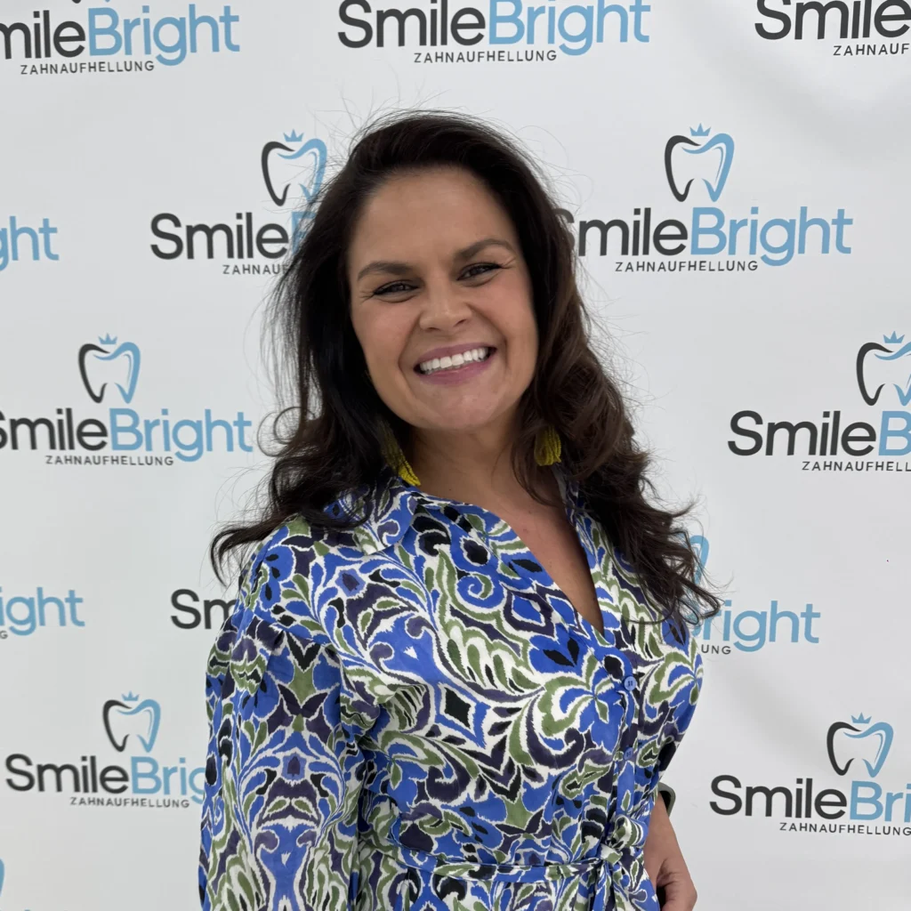 Eine strahlend lächelnde Frau mit lockigem Haar in einem gemusterten Kleid posiert vor der smileBright Logo-Wand für Zahnaufhellung in Düsseldorf.